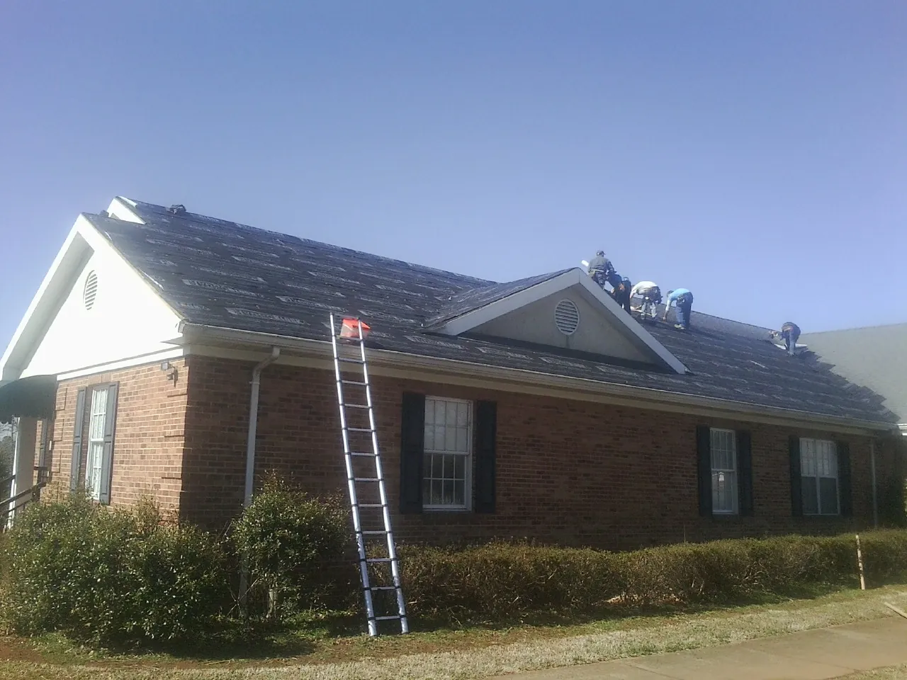 Crew tearing shingles off a brick ranch roof with a ladder set — Metal Roof Repair work by our crew in Tupelo