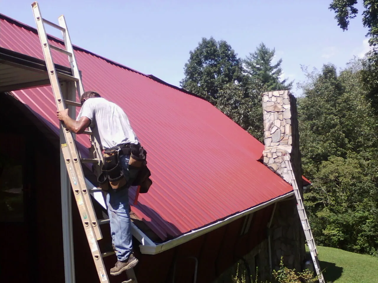 Roofer on a ladder beside a red metal roof and stone chimney — Soffit & Fascia Repair by Roofing Brothers in Tupelo